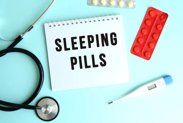 The text SLEEPING PILLS is written on a white notepad that lies next to the stethoscope and pills on a blue background. Medical concept