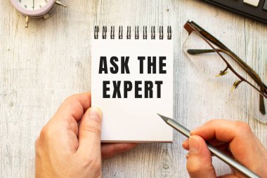 The hand writes the text ASK THE EXPERT in pencil in a notebook. View from above. Business concept.