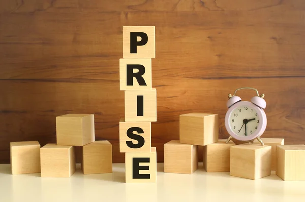 Five wooden cubes stacked vertically on a brown background make up the word PRISE. Cubes are scattered nearby and there is a clock. Front View Concepts