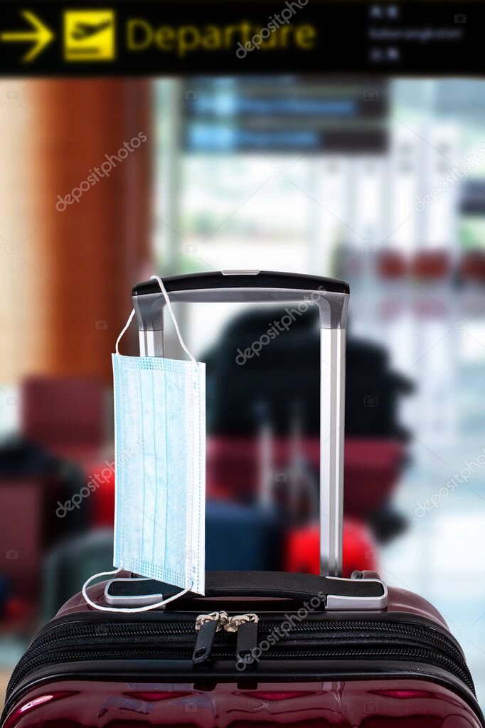 Travel suitcase and medical mask with defocused airport background. Concept of travel restrictions or ban during Covid-19 pandemic and quarantine against coronavirus. Vertical orientation.
