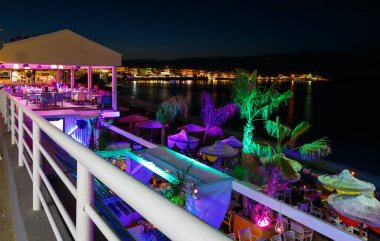 Gece sahillerindeki korkuluk ve rıhtım kafeleri boyunca bir plaj ve şemsiyeyle manzara ve parlak renklerle vurgulanmış Girit, Yunanistan