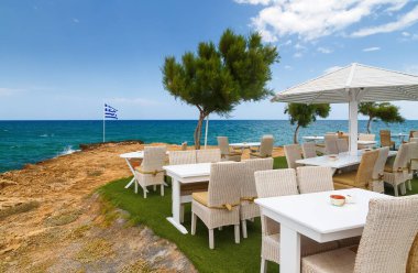 Denize bakan kayalık sahilde masa sandalyeleri ve ahşap şemsiye ve Yunan bayrağı Girit, Yunanistan