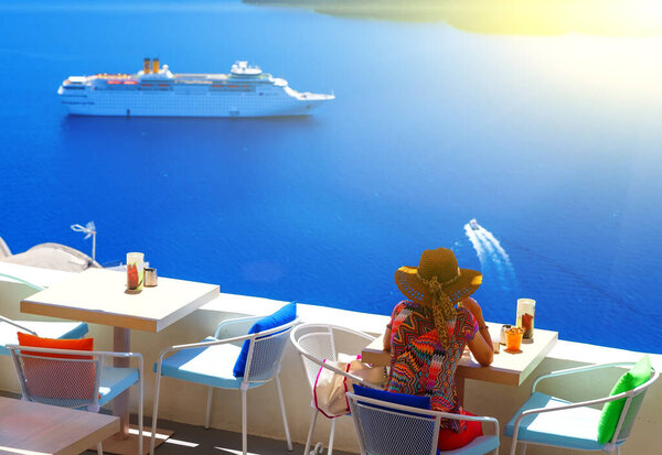 woman in hat sitting in a cafe and enjoys views of the Caldera and cruise ship, Santorini, Fira, Greece