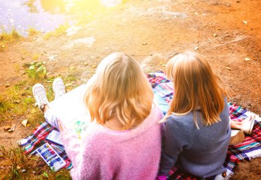 Parktaki gölün kıyısında iki genç sarışın oturuyor ve resim çiziyor.