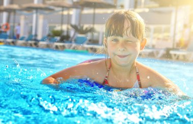 Gülümseyen küçük kız havuzda göz kırpıyor. Çocuk kaydırağı ve arka planda güneş ışınları