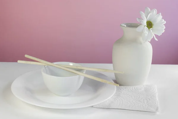 Minimalist still life in oriental style with  white vase, chrysanthemum, plate, cup, chopsticks and napkin on a pink background
