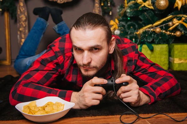 A man with a beard and mustache in a red checkered shirt is playing with a joystick. Eating chips. Against the background of the New Year tree. New year decoration. Holiday weekend