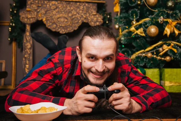 A man with a beard and mustache in a red checkered shirt is playing with a joystick. Eating chips. Against the background of the New Year tree. New year decoration. Holiday weekend