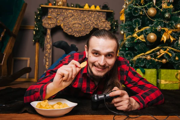 一个留着胡子和胡子 穿着红色格子衬衫的男人在玩操纵杆 吃薯片在新年树的背景下 新年装饰 假日周末 — 图库照片