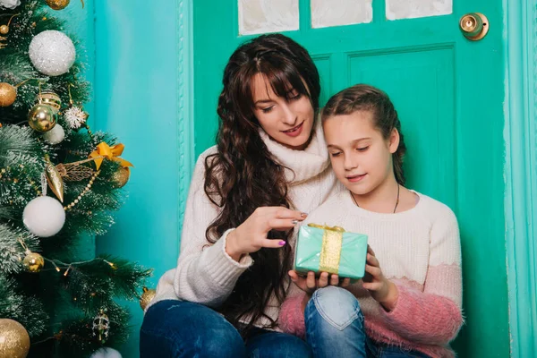 Mom and daughter are sitting on the steps near the Christmas door and waiting for the new year. A loving family, they cut the Christmas tree and give gifts. Woman and girl eight years old in the scenery