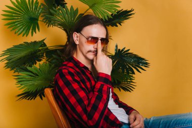 80 'lerin kırmızı disko tişörtlü, uzun saçlı ve bıyıklı çekici bir adam sarı arka planda bir sandalyede oturuyor. Yeşil palmiye ağacının arka planında gözlüklü bir adam.