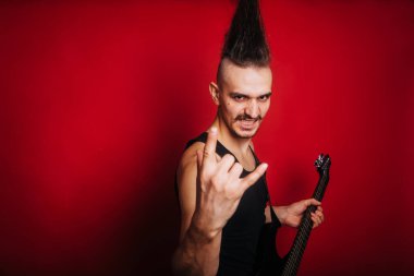 Punk with black electric guitar, mustache and big mohawk showing horn gesture. Photo of a rock musician in the studio on a red background. Informal movement clipart