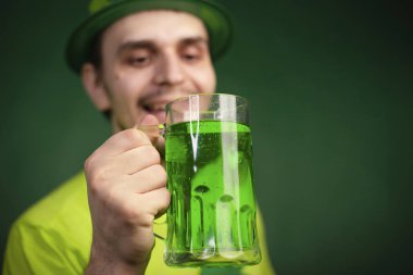 Yonca şapkalı ve yeşil tişörtlü bir adam büyük bir bardak yeşil bira içiyor. Adam Aziz Patrick gününü bir bardak birayla kutluyor..