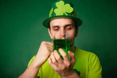 Yonca şapkalı ve yeşil tişörtlü bir adam büyük bir bardak yeşil bira içiyor. Adam Aziz Patrick gününü bir bardak birayla kutluyor..