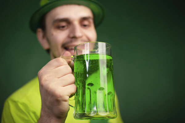 Мужчина в шляпе и зеленой футболке пьет большую кружку зеленого эля. Парень празднует День Святого Патрика с бокалом пива.