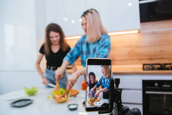 Two girls shoot a video on a smartphone as they cook in the kitchen. Women bloggers shoot the recipe