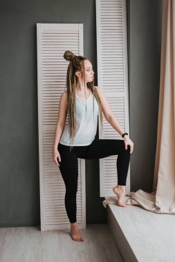 Spor kıyafetleri içinde uzun örgülü genç bir kadın yoga yapıyor. Güzel bir kız büyük bir pencerenin yanında duruyor. Büyük stüdyo salonu