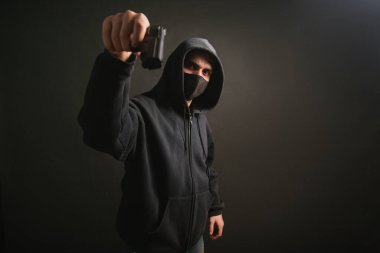 Kapüşonlu, maskeli bir gangster karanlık bir arka planda elinde silah tutuyor. Haydut tabancayla nişan alıyor. Fotoğraf stüdyoda..