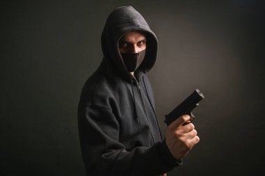 Kapüşonlu, maskeli bir gangster karanlık bir arka planda elinde silah tutuyor. Haydut tabancayla nişan alıyor. Fotoğraf stüdyoda..