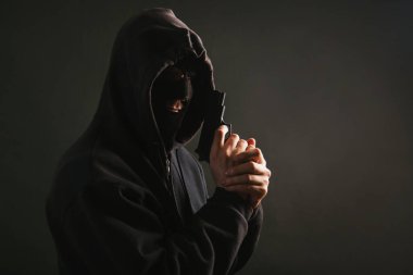 Kapüşonlu, maskeli bir gangster karanlık bir arka planda elinde silah tutuyor. Haydut tabancayla nişan alıyor. Fotoğraf stüdyoda..
