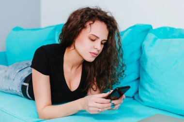 Genç ve güzel bir kadın elinde bir akıllı telefon tutuyor, yazı yazıyor, mavi bir kanepede yatıyor, cep telefonuyla çalışıyor. İnternet aramaları