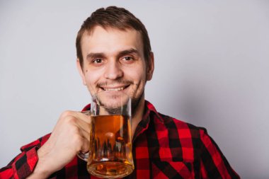Kırmızı tişörtlü genç adam beyaz arka planda köpüksüz büyük bir bardak bira tutuyor. Kötü alkol içiyor.
