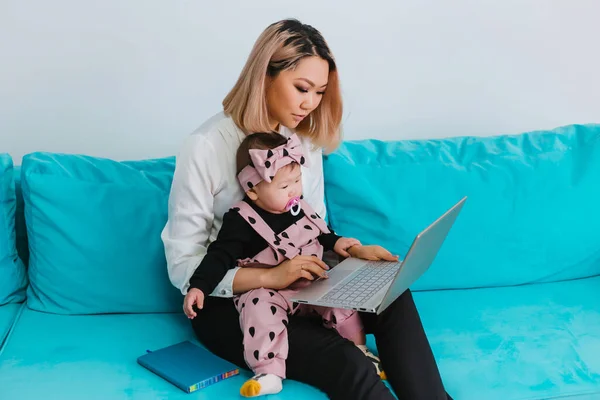 A young mother works over the Internet. A woman of Asian appearance uses a laptop, holding a baby in her arms, sitting on a blue sofa. Work from home