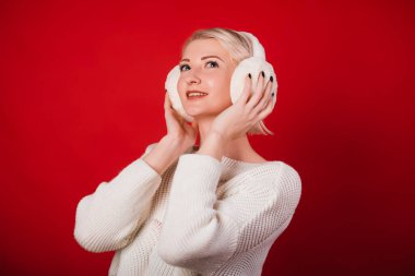 Beyaz örgü süveter ve kürk kulaklıklı genç bir kadın kırmızı arka planda duruyor. Kış kıyafetleri içinde tatlı bir gülümsemesi olan bir kız. Stüdyo fotoğrafı