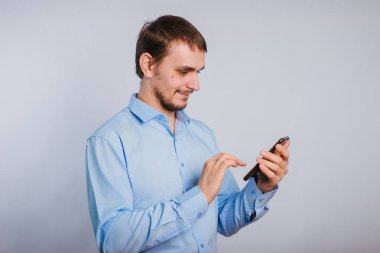 Mavi gömlekli bir adam beyaz arka planda akıllı telefon tutuyor. Erkek bir ofis çalışanı telefonda yazı yazıyor..