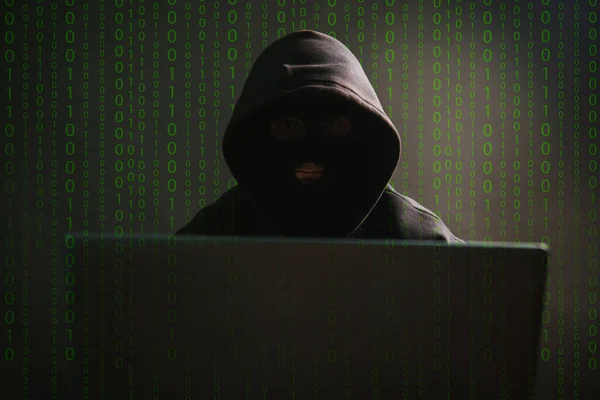 A dangerous masked hooded hacker infiltrates data servers and infects their systems with viruses. Male criminal steals information through a laptop. Photo on a dark background in the studio