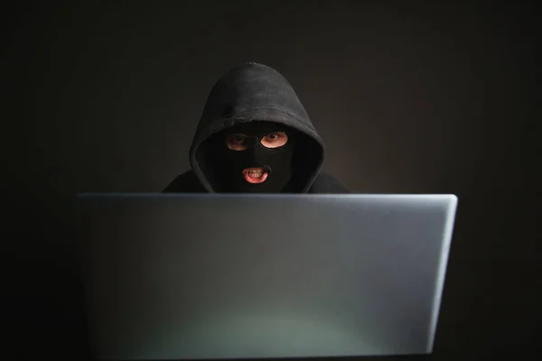 An angry hacker in a mask with a hood infiltrates data servers and infects their systems with viruses. A male criminal steals information through a laptop and is defeated. Photo on a dark background in the studio