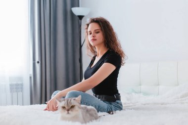 Genç ve güzel bir kız beyaz bir odada elinde akıllı bir telefonla bir yatakta oturuyor. Yakınlarda bir İskoç gri kedisi yatıyor. Ev konforu