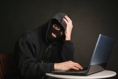 Kapüşonlu tehlikeli maskeli bir hacker veri sunucularına sızıp onlara virüs bulaştıramaz. Suçluyu bir dizüstü bilgisayarla yakaladık. Stüdyoda karanlık bir arkaplanda fotoğraf.