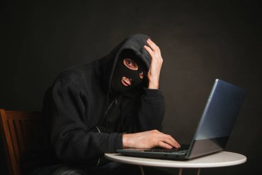 Kapüşonlu tehlikeli maskeli bir hacker veri sunucularına sızıp onlara virüs bulaştıramaz. Suçluyu bir dizüstü bilgisayarla yakaladık. Stüdyoda karanlık bir arkaplanda fotoğraf.