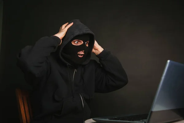 A dangerous masked hacker with a hood cannot infiltrate the data servers and infect them with viruses. Caught the criminal through a laptop. Photo on a dark background in the studio