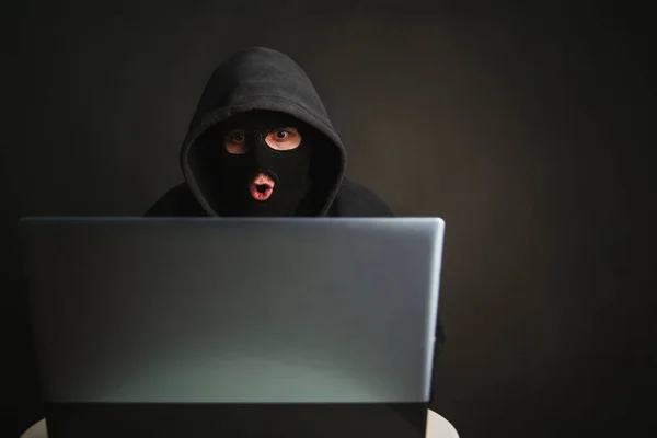 An angry hacker in a mask with a hood infiltrates data servers and infects their systems with viruses. A male criminal steals information through a laptop and is defeated. Photo on a dark background in the studio