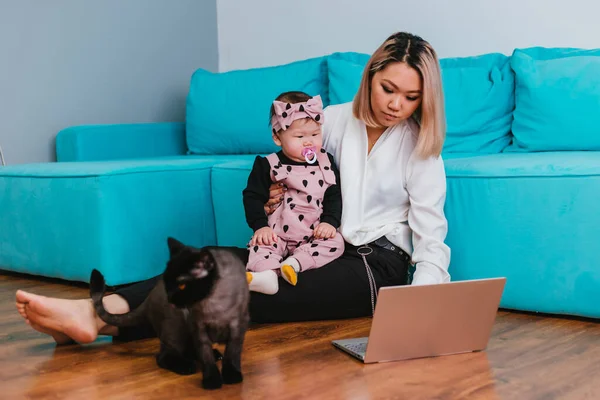 若い母親はインターネット上で働いている アジア系の女性は 猫の横の床に座って 彼女の腕の中で赤ちゃんを保持し ラップトップを使用しています 家からの仕事 — ストック写真