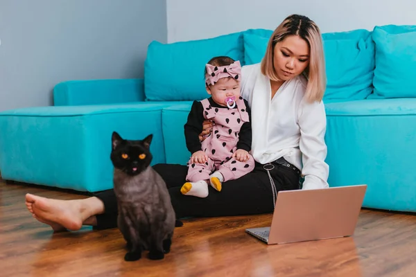 若い母親はインターネット上で働いている アジア系の女性は 猫の横の床に座って 彼女の腕の中で赤ちゃんを保持し ラップトップを使用しています 家からの仕事 — ストック写真