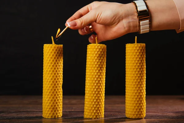 A hand lights a beeswax candle stands on the table. Eco product on a dark background