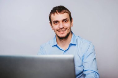 Mavi gömlekli genç yakışıklı bir adam bilgisayarın başında oturuyor. Beyaz arka planda çalışan bir ofis çalışanı bir dizüstü bilgisayar için çalışır. izole et