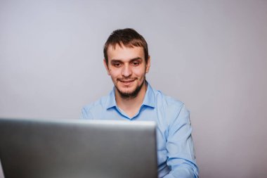 Mavi gömlekli genç yakışıklı bir adam bilgisayarın başında oturuyor. Beyaz arka planda çalışan bir ofis çalışanı bir dizüstü bilgisayar için çalışır. izole et