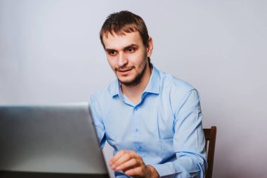 Mavi gömlekli genç yakışıklı bir adam bilgisayarın başında oturuyor. Beyaz arka planda çalışan bir ofis çalışanı bir dizüstü bilgisayar için çalışır. izole et