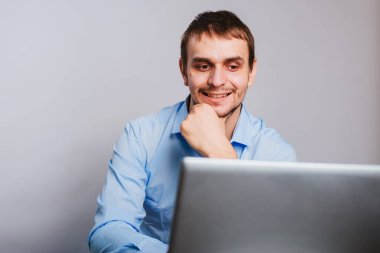 Mavi gömlekli genç yakışıklı bir adam bilgisayarın başında oturuyor. Beyaz arka planda çalışan bir ofis çalışanı bir dizüstü bilgisayar için çalışır. izole et