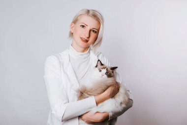 Beyaz önlüklü bir kız tüylü bir Regg Dol kedisi tutuyor. Veterenar hayvanı tarıyor. Hayvan bakımı