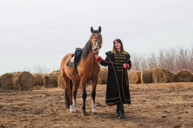 Tarihi süvari kostümü içinde uzun ve siyah saçlı güzel bir kadın bir at ile bir tarlada duruyor. At tutan bir kız