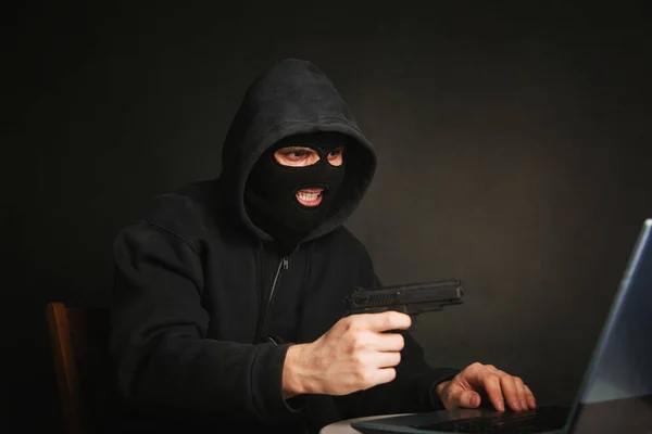 黒いマスクの犯罪者とピストルを持ったフードはノートパソコンを通してお金を盗む ハッカーの攻撃だ オンライン盗難の概念 — ストック写真