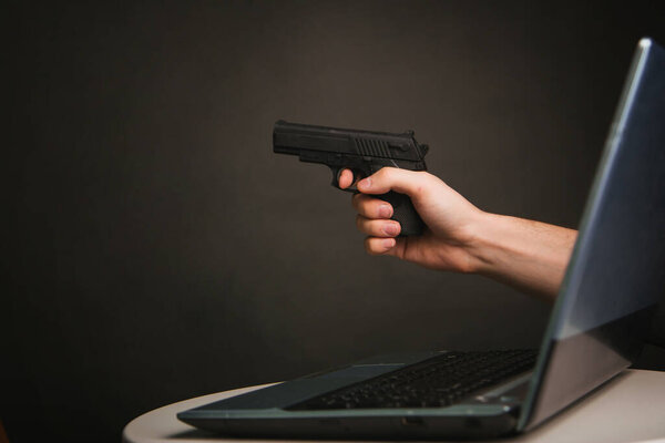 рука с пистолетом из ноутбука на темно-сером фоне. Концепция кражи онлайн