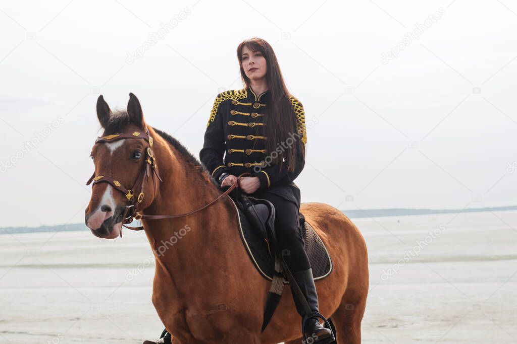 A beautiful woman with long and black hair in a historical hussar costume stands near a river with a horse. Girl holding horse