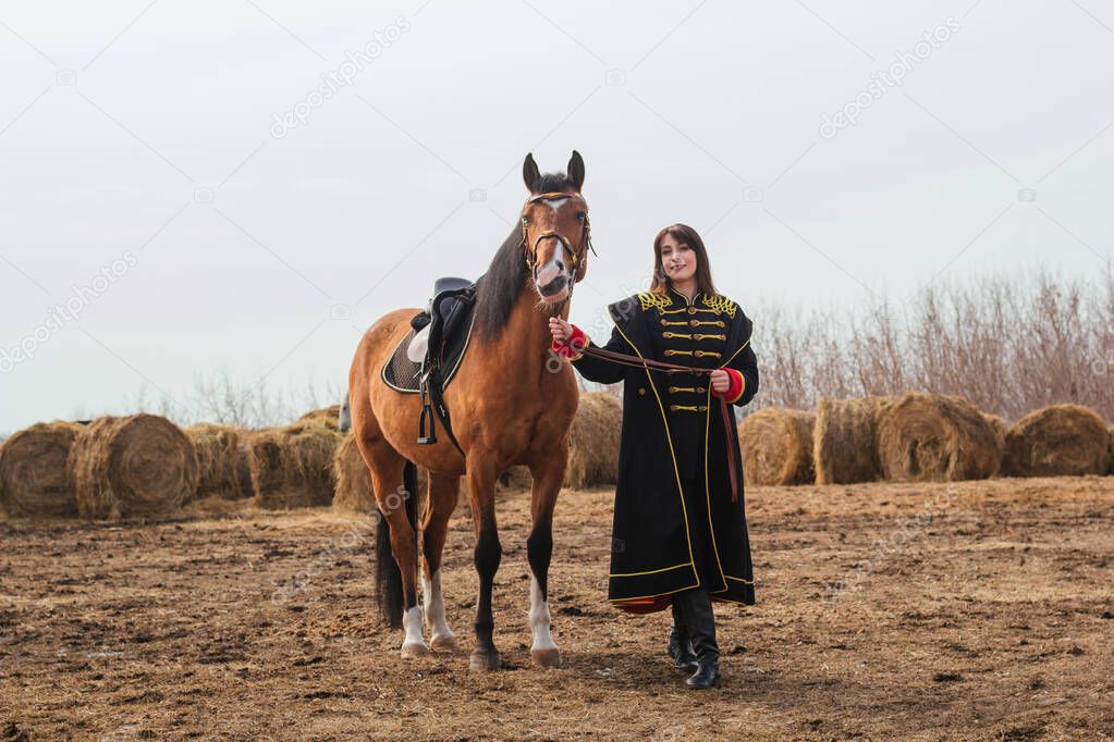 A beautiful woman with long and black hair in a historical hussar costume stands in a field with a horse. A girl holding a horse