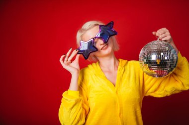 Mor büyük gözlüklü, yıldız şeklinde ve sarı pijamalı parlak bir kız. Elinde disko topu olan komik ve neşeli bir kadın. Kırmızı arkaplanda stüdyo fotoğrafı